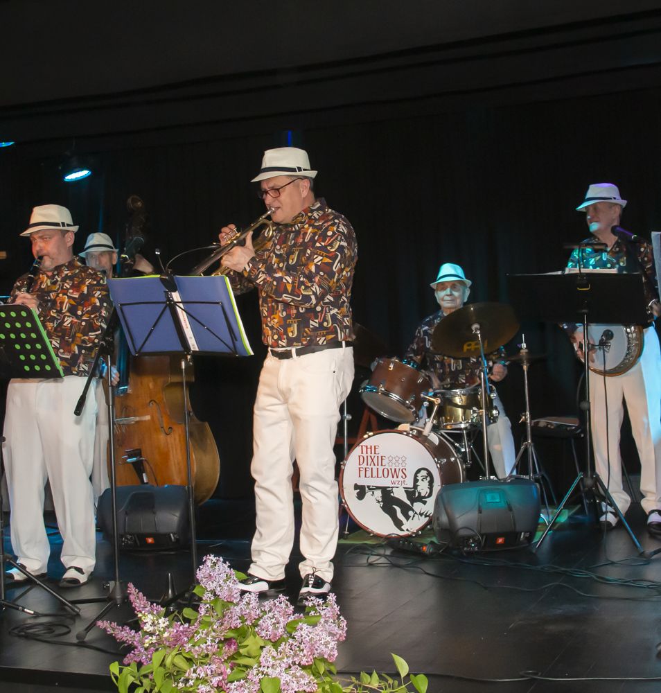 Elegancko ubrani Panowie grają na scenie na instrumentach typowo jazzowych (trąbka, kontrabas, puzon, gitara itp). Pod sceną tańczą i uśmiechają się uczestnicy koncertu połączonego z potańcówką.