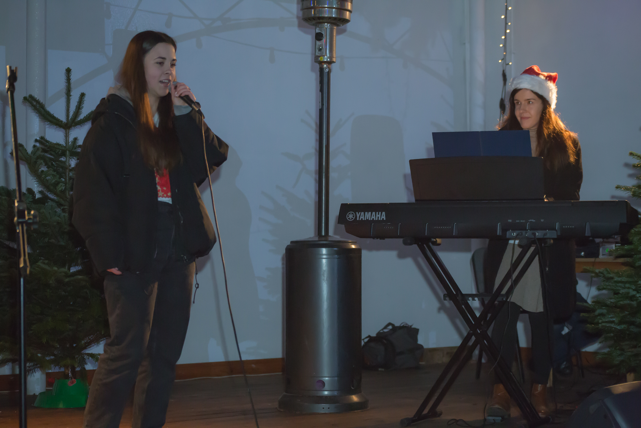 Na scenie młoda dziewczyna śpiewa do mikrofonu. Druga młoda kobieta w czapce świątecznej gra na elektrycznym pianinie. Obie panie są rozpromienione. W tle dekoracja świąteczna, żywe choinki, światełka choinkowe oraz latarnia ogrzewająca scenę.