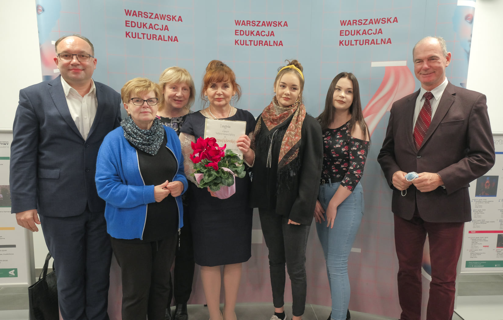 Na tle ścianki prasowej stoi dyrektor Domu Kultury Rembertów, nagrodzona Marzenna Kosowska z dyplomem i kwiatami oraz Zastępca Burmistrza Dzielnicy Rembertów