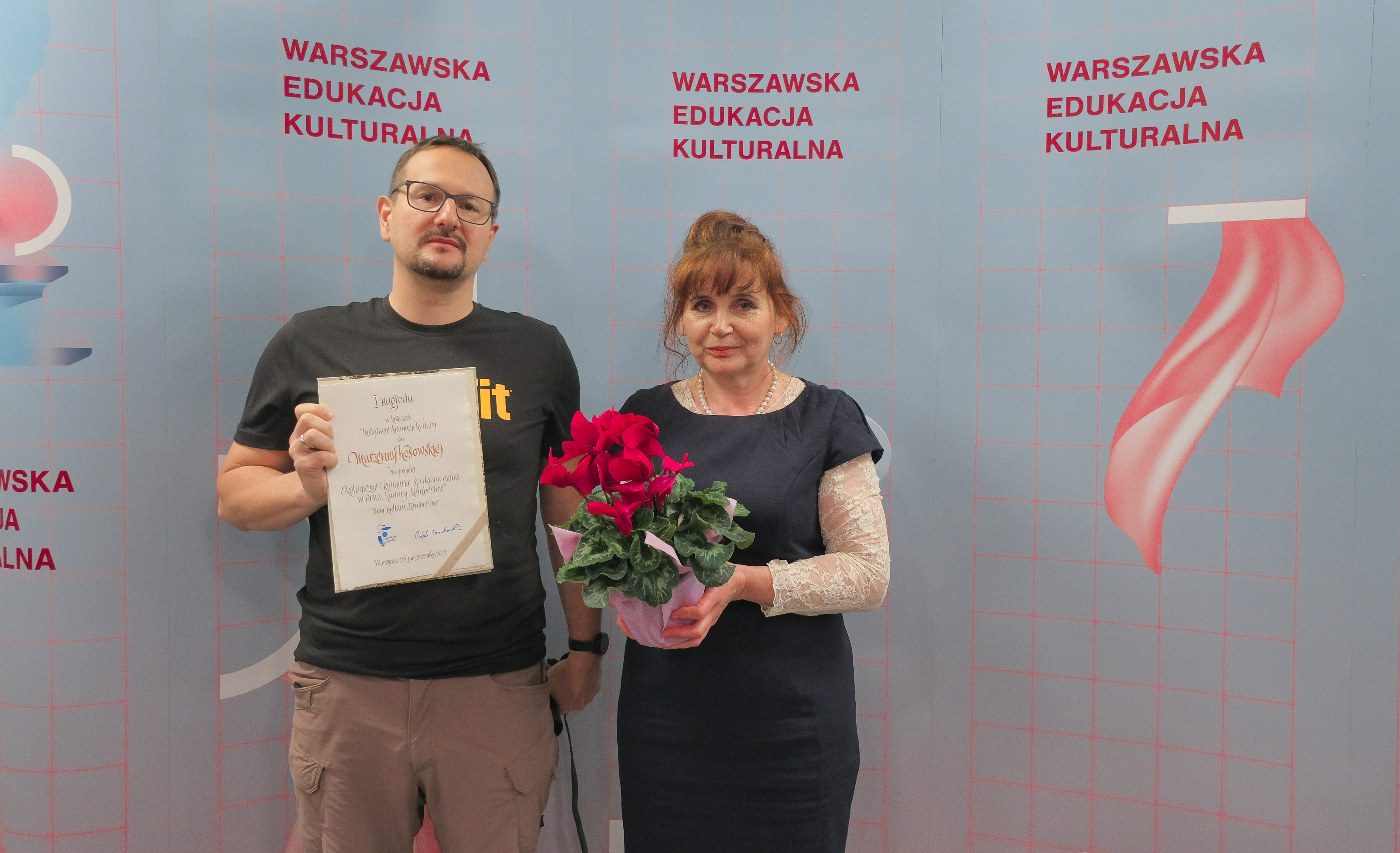 Na tle ścianki prasowej z dyplomem stoi Marcin Sochacki oraz pani Marzenna z kwiatami