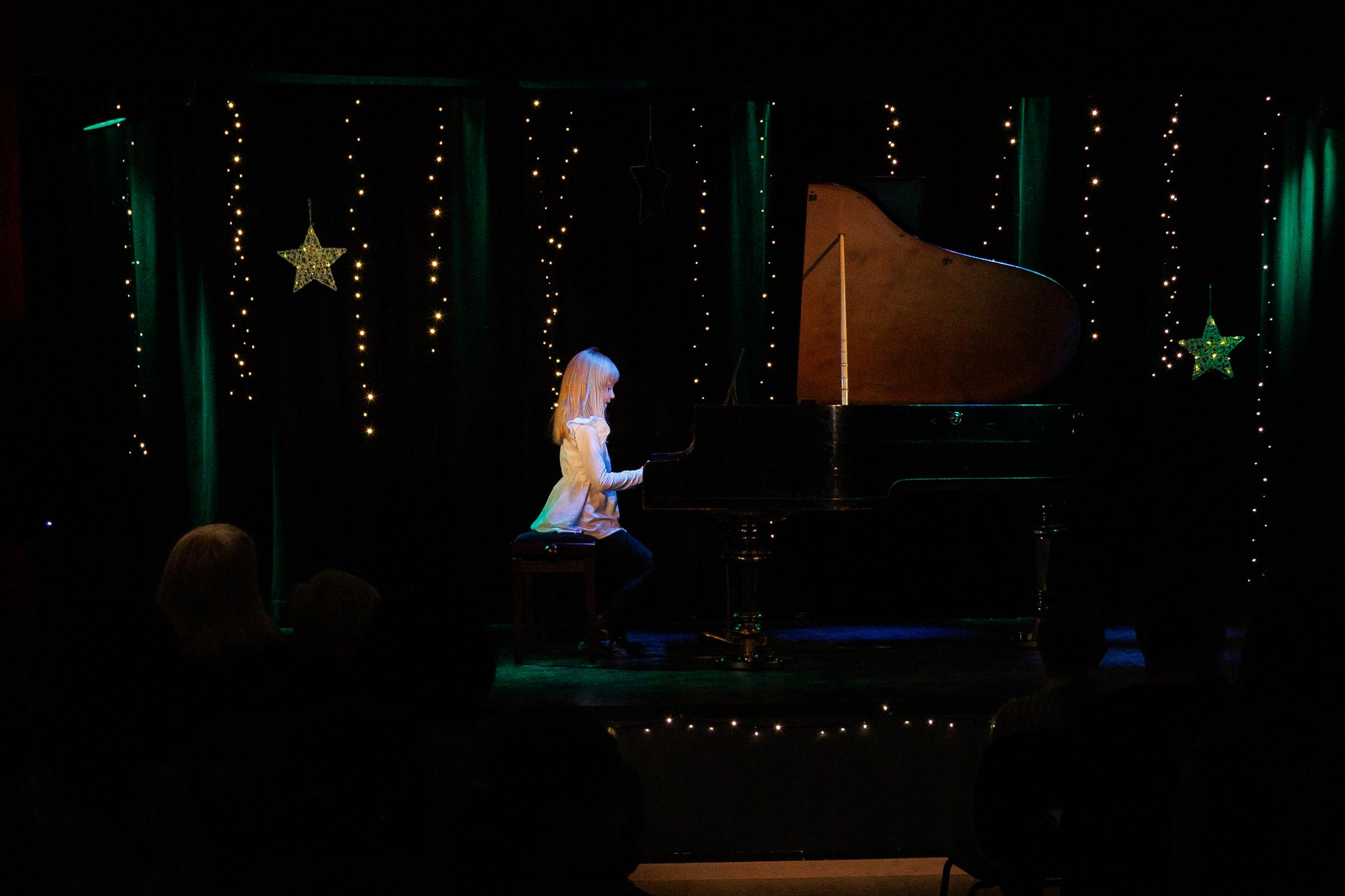 scena, fortepian, mała dziewczynka gra na fortepianie, ma ładną jasna sukienkę, włosy ładnie zaczesane, blondynka. Gra w wielkim skupieniu.