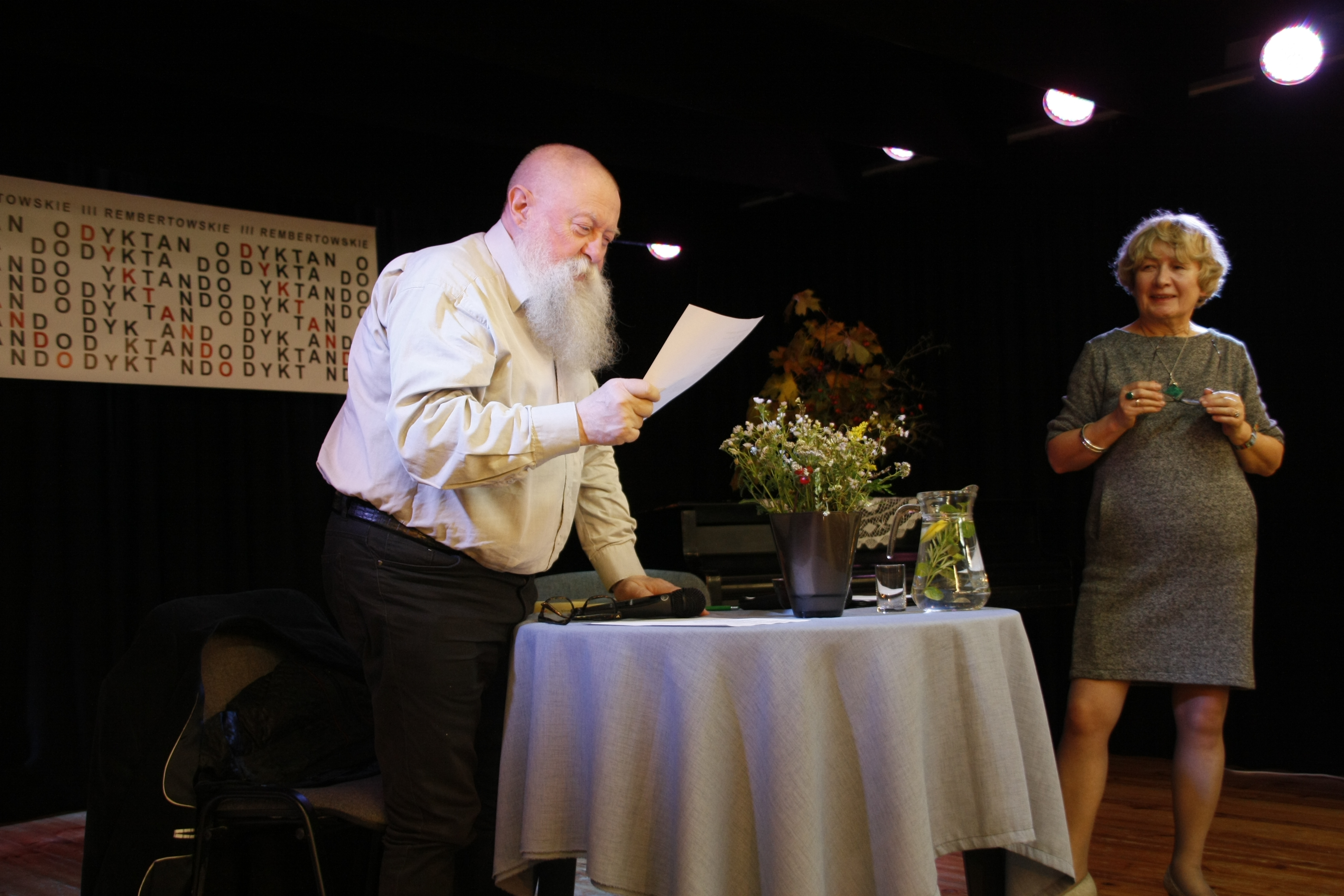 Na scenie stoi profesor Bralczyk odczytujący treść dyktanda, opiera się o przystrojony kwiatami stolik. Uważnie słucha go doktor Majkowska
