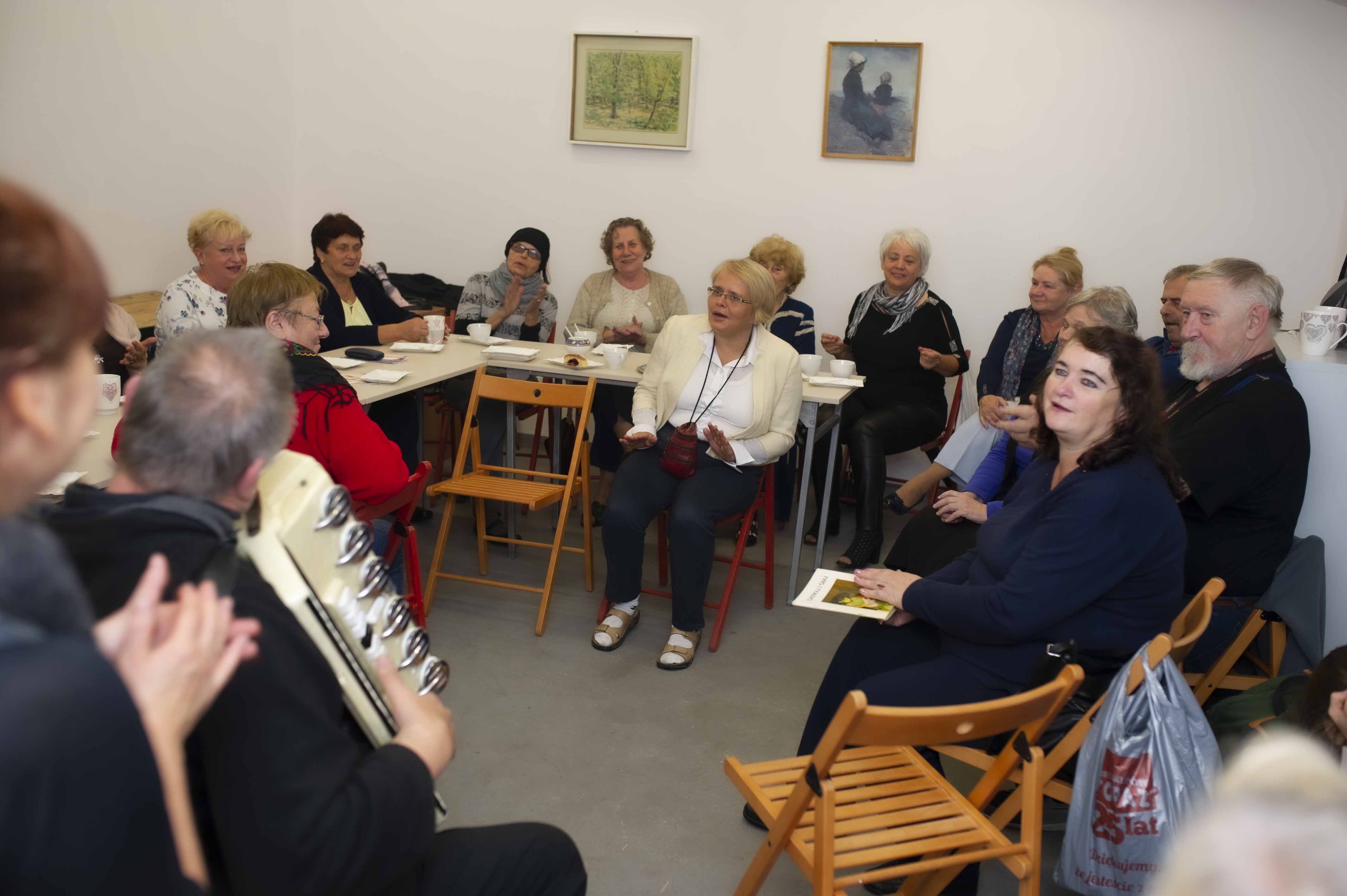 Podwieczorek przy akordeonie w Szopie Dobrych Pomysłów działającej przy domu kultury. Zdjęcie przedstawia grupę śpiewających seniorów, którzy siedzą dookoła pomieszczenia. Po lewej stronie zdjęcia tyłem do aparatu, a przodem do publiczności siedzi muzyk grający na akordeonie. Panuje radosna atmosfera.