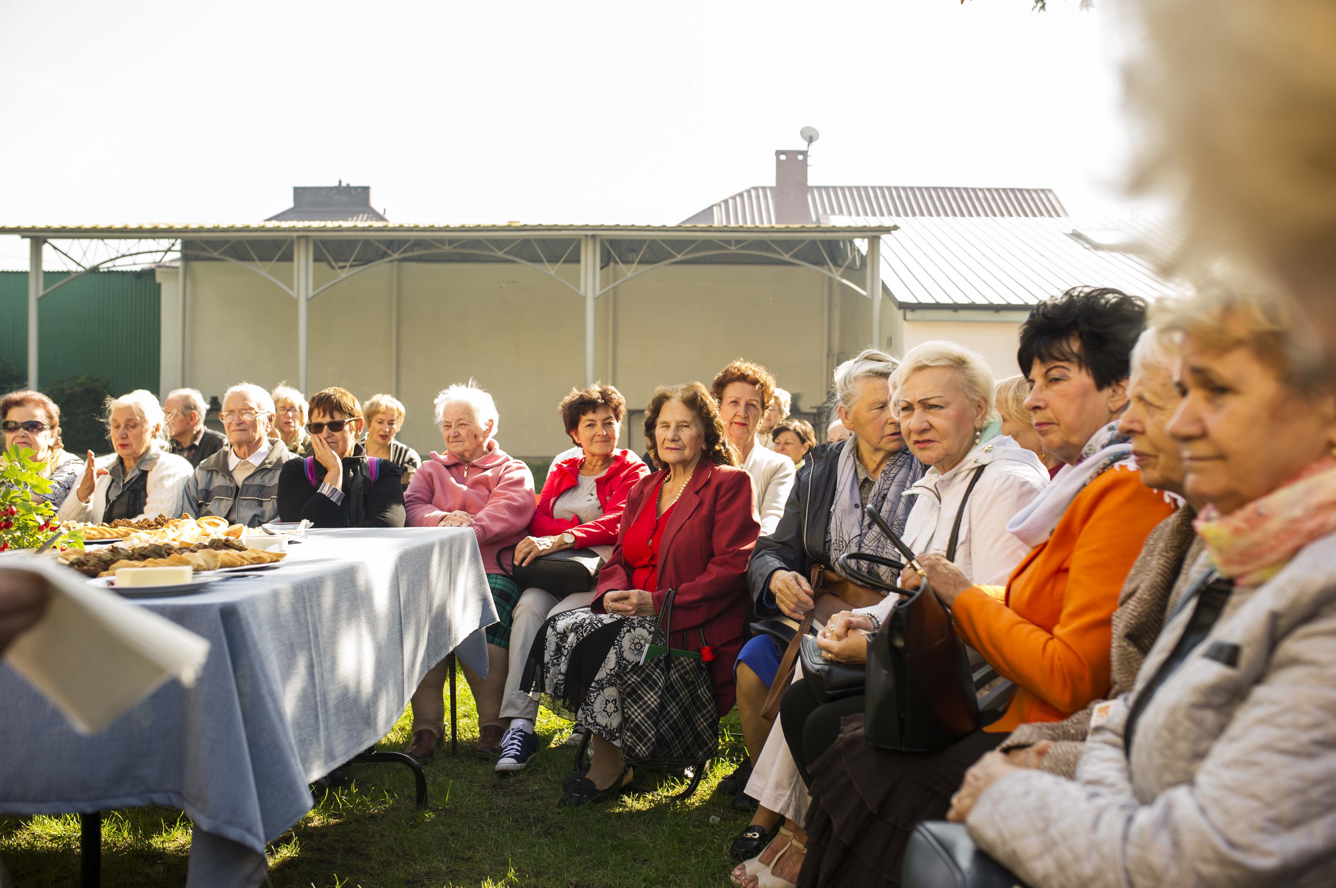 Śniadanie francuskie dla seniorów. Na słonecznym dziedzińcu domu kultury przy zastawionym francuskimi smakołykami stole siedzą zgromadzeni seniorzy.