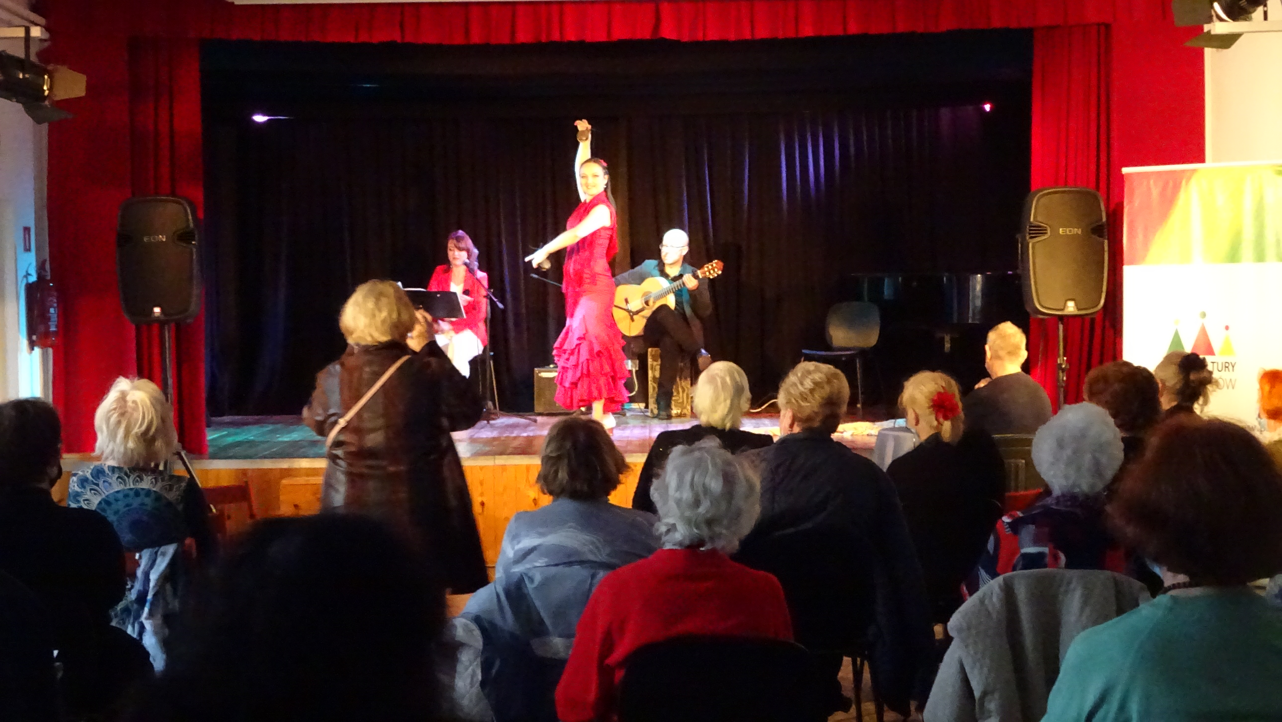 Wieczór Hiszpański dla seniorów. Przed sceną widzimy publiczność trwa pokaz tańca Flamenco. Na scenie w czerwonej sukni, z uniesioną jedną ręką, z drugą ręką przed sobą zastygła tancerka. Za nią siedzi wokalistka oraz gitarzysta.